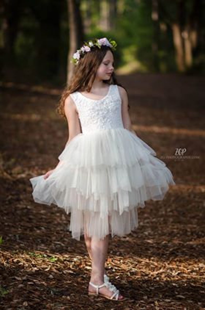 May include: A young girl wearing a white tulle dress with a floral crown. The dress has multiple layers and a high neckline. The girl is standing in a wooded area.