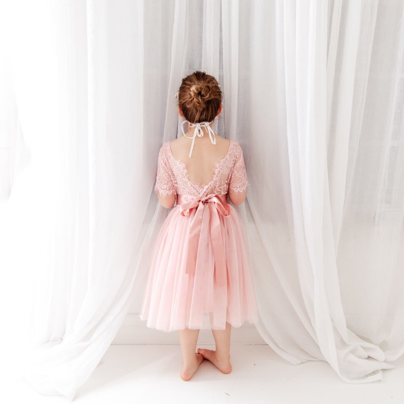 Pode incluir: Uma menina pequena vestindo um vestido de tule rosa com detalhes em renda e uma faixa de cetim rosa combinando. O vestido tem costas baixas e &eacute; usado com uma fita branca ao redor do pesco&ccedil;o.
