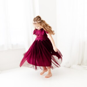 May include: A young girl wearing a burgundy tulle dress with a lace bodice and a gold crown. She is twirling and smiling.
