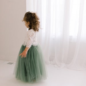 May include: A young girl wearing a white lace top and a sage green tulle skirt. The skirt is full and flowing, and the girl is standing in front of a white curtain.