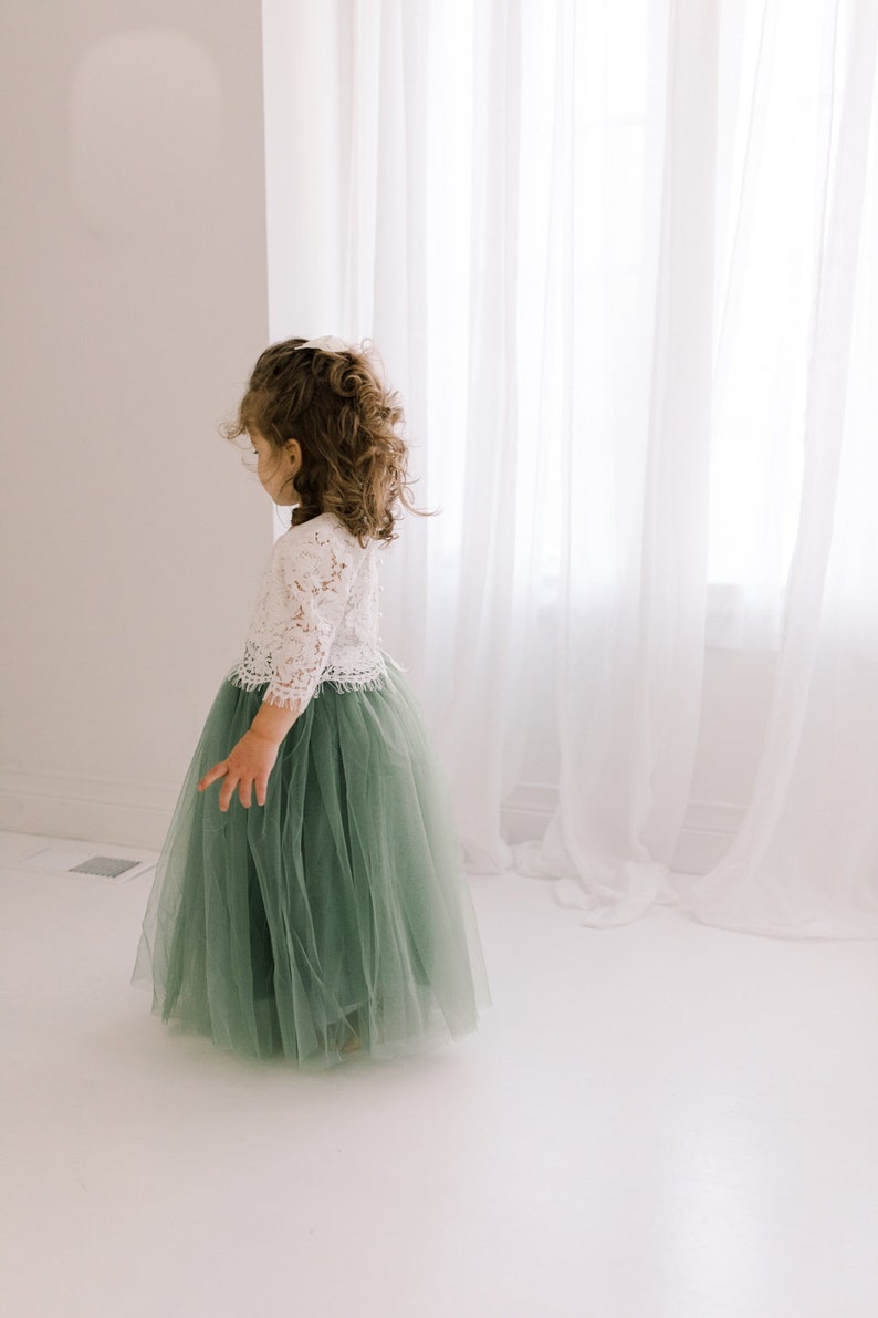 May include: A young girl wearing a white lace top and a sage green tulle skirt. The skirt is full and flowing, and the girl is standing in front of a white curtain.