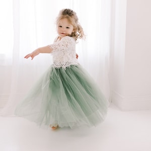 May include: A young girl wearing a white lace top and a sage green tulle skirt. She is twirling and smiling at the camera.