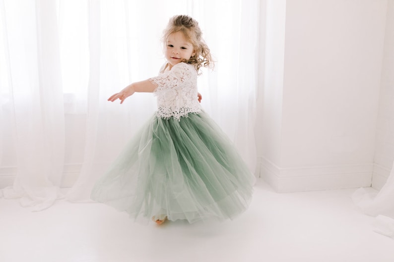 May include: A young girl wearing a white lace top and a sage green tulle skirt. She is twirling and smiling at the camera.