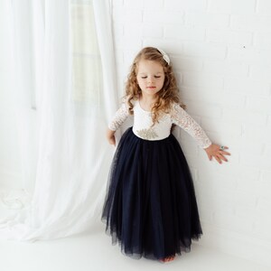 May include: A young girl wearing a white lace long-sleeved top with a navy blue tulle skirt. She is standing in front of a white brick wall.