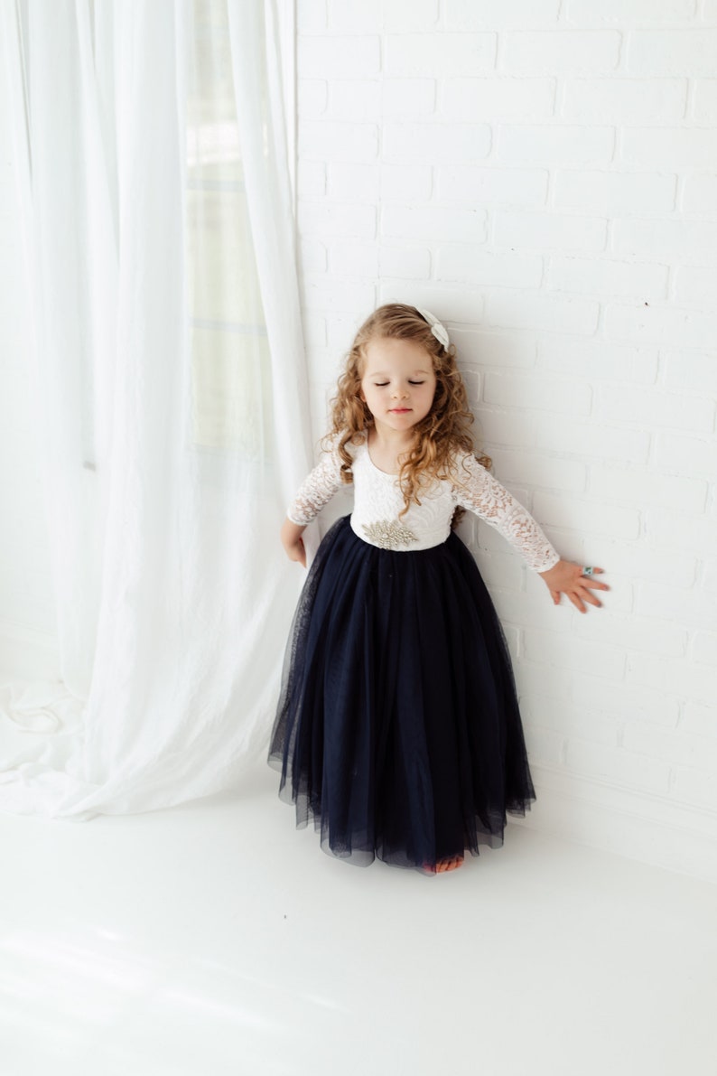 May include: A young girl wearing a white lace long-sleeved top with a navy blue tulle skirt. She is standing in front of a white brick wall.