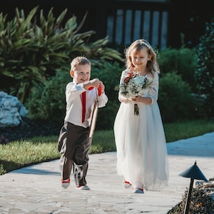 Puede incluir: Una ni&ntilde;a peque&ntilde;a con un vestido blanco y un ni&ntilde;o peque&ntilde;o con una camisa blanca y pantalones grises caminan por un camino de piedra. La ni&ntilde;a lleva un ramo de flores blancas y rojas. El ni&ntilde;o lleva un cartel de madera.