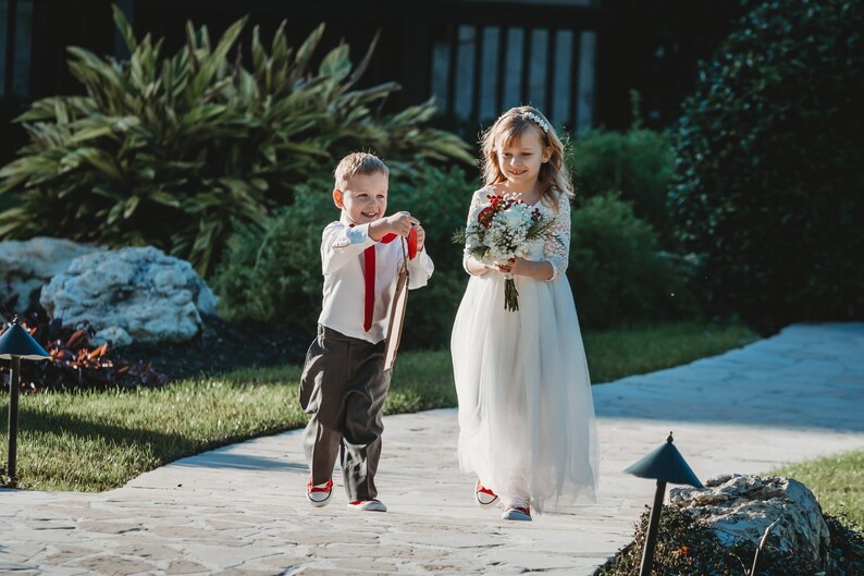 Puede incluir: Una ni&ntilde;a peque&ntilde;a con un vestido blanco y un ni&ntilde;o peque&ntilde;o con una camisa blanca y pantalones grises caminan por un camino de piedra. La ni&ntilde;a lleva un ramo de flores blancas y rojas. El ni&ntilde;o lleva un cartel de madera.