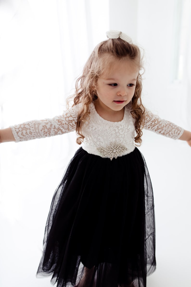 May include: A dress featuring a white lace bodice with three-quarter length sleeves and a full black tulle skirt. The waist is accented with a decorative silver-toned brooch made of clear crystals. A small white pearl-adorned hair accessory is visible.