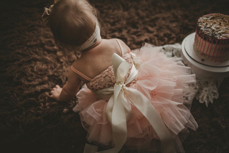 May include: A young child wearing a pink tulle dress with a white satin bow. The dress has a lace bodice and the child is sitting on a brown carpet.