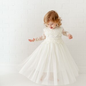 May include: A floor-length cream-colored dress featuring long sheer lace sleeves, a high neckline, and a bodice adorned with intricate floral lace overlays. The skirt is constructed from multiple layers of soft tulle, creating a full silhouette.