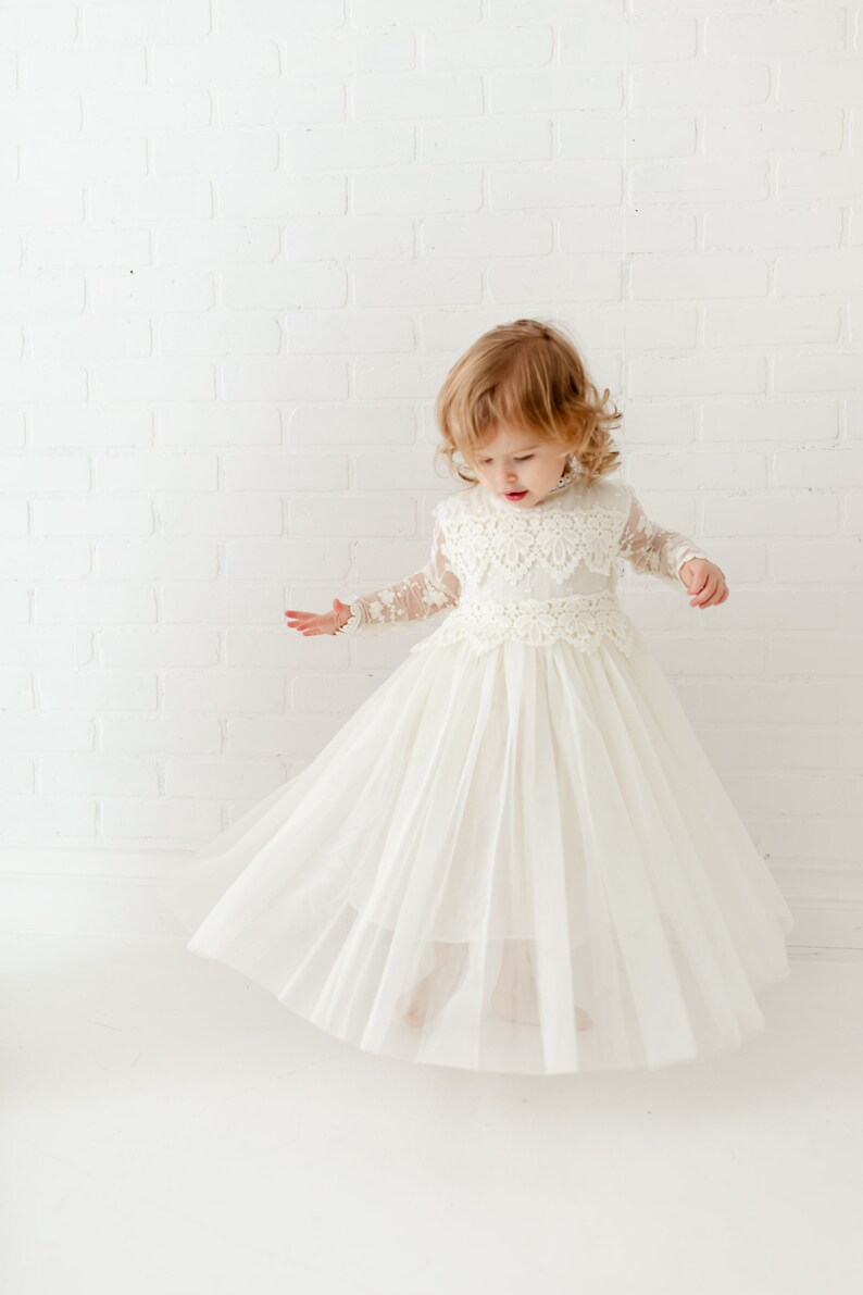 May include: A floor-length cream-colored dress featuring long sheer lace sleeves, a high neckline, and a bodice adorned with intricate floral lace overlays. The skirt is constructed from multiple layers of soft tulle, creating a full silhouette.