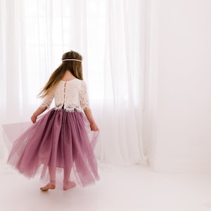 May include: A young girl wearing a white lace top and a long, flowing mauve tulle skirt. She is standing in front of a white window with sheer curtains.