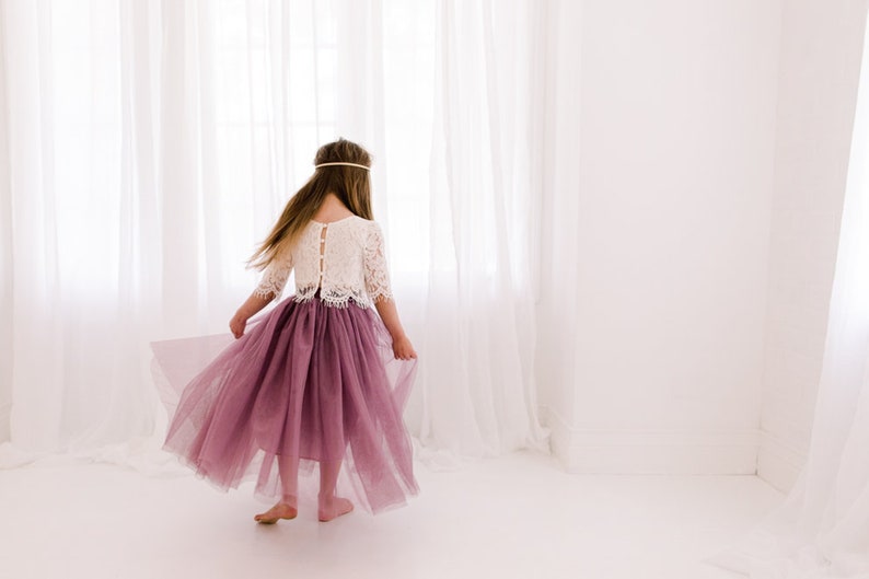 May include: A young girl wearing a white lace top and a long, flowing mauve tulle skirt. She is standing in front of a white window with sheer curtains.