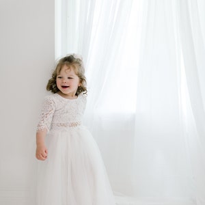 May include: A young girl in a white lace top and tulle skirt stands in front of a white window with sheer curtains. She is smiling and looking to the side.