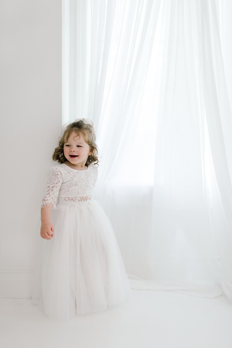 May include: A young girl in a white lace top and tulle skirt stands in front of a white window with sheer curtains. She is smiling and looking to the side.