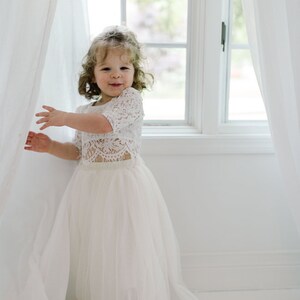 May include: A young girl in a white lace crop top and a long, flowing tulle skirt. The outfit is a light ivory color. She is standing near a window with white curtains, smiling at the camera.