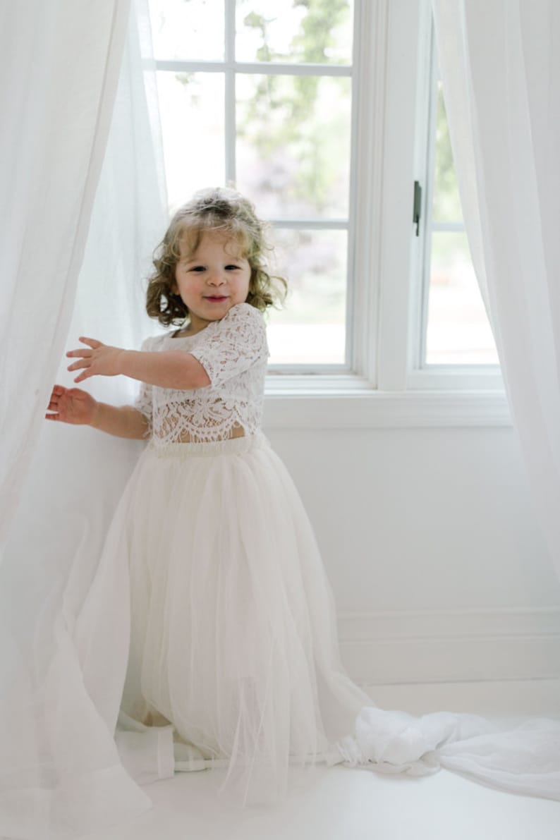 May include: A young girl in a white lace crop top and a long, flowing tulle skirt. The outfit is a light ivory color. She is standing near a window with white curtains, smiling at the camera.