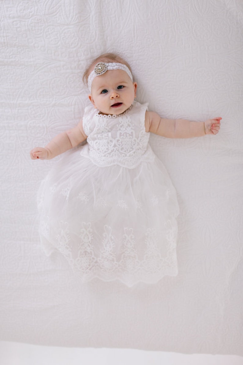 May include: A baby girl wearing a white lace dress and a white headband with a jeweled accent. The baby is lying on a white blanket.