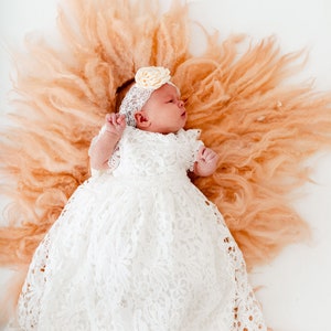 May include: A baby girl wearing a white lace dress and a flower headband lies on a peach-colored fluffy blanket.