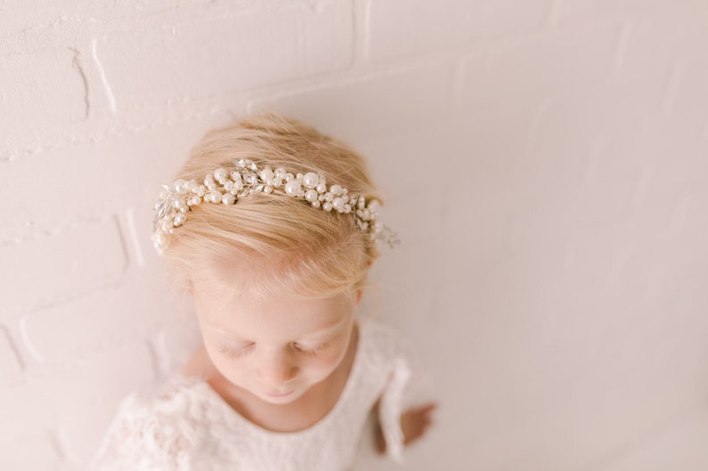 May include: A young girl wearing a white dress with a pearl and crystal hairpiece. The hairpiece is a delicate headband with small pearls and rhinestones.
