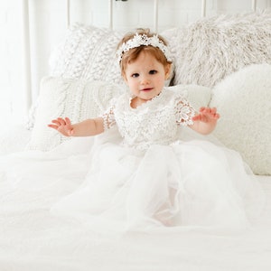 May include: A white dress featuring a lace bodice with short sleeves and a layered tulle skirt. The garment is paired with a matching white lace headband. The design includes intricate floral patterns on the lace bodice and sleeves.