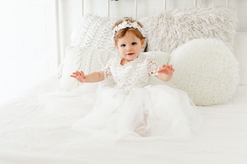 May include: A white dress featuring a lace bodice with short sleeves and a layered tulle skirt. The garment is paired with a matching white lace headband. The design includes intricate floral patterns on the lace bodice and sleeves.