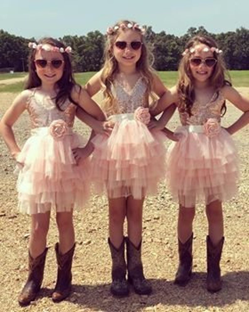 以下が含まれることがあります： 花冠とサングラスをつけた3人の女の子が、ピンクと白のキラキラしたチュールスカートのドレス、花のサッシュ、カウボーイブーツを履いています。