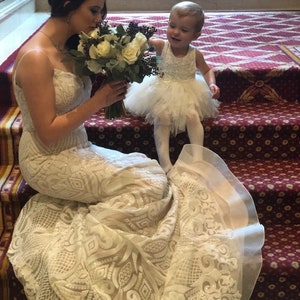 K&ouml;nnte beinhalten: Eine Braut in einem wei&szlig;en Spitzen-Hochzeitskleid mit langem Schleier, die einen Strau&szlig; wei&szlig;er Blumen h&auml;lt. Ein junges M&auml;dchen in einem wei&szlig;en T&uuml;llkleid steht neben ihr und l&auml;chelt.