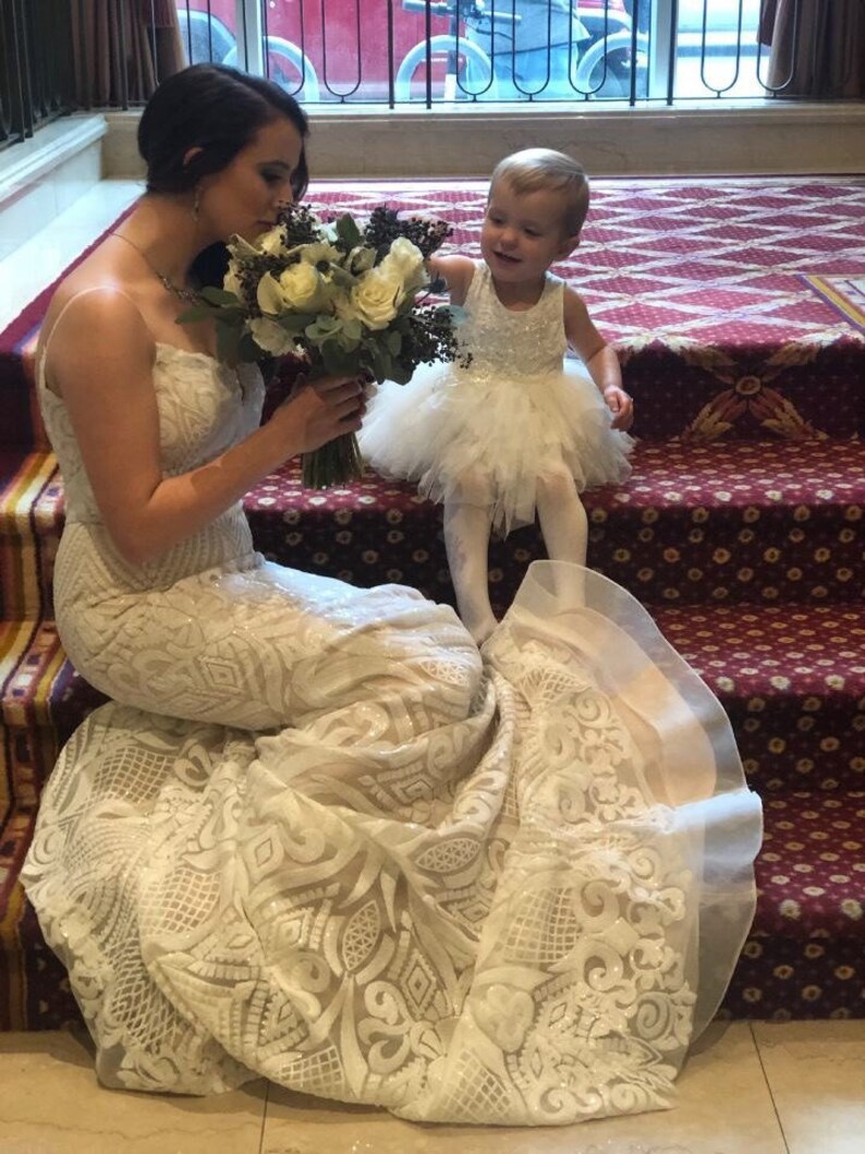 K&ouml;nnte beinhalten: Eine Braut in einem wei&szlig;en Spitzen-Hochzeitskleid mit langem Schleier, die einen Strau&szlig; wei&szlig;er Blumen h&auml;lt. Ein junges M&auml;dchen in einem wei&szlig;en T&uuml;llkleid steht neben ihr und l&auml;chelt.