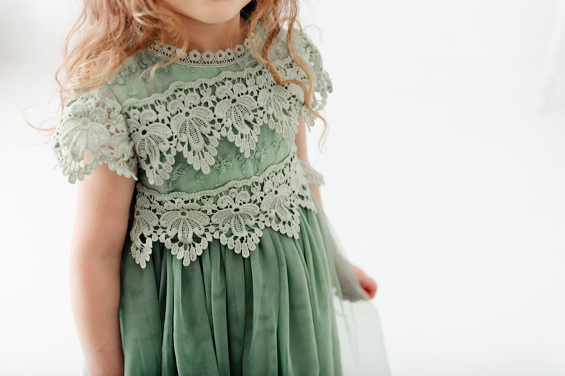 May include: A sage green tulle dress with delicate lace detailing around the neckline and bodice. The dress has a flowing skirt and short sleeves.