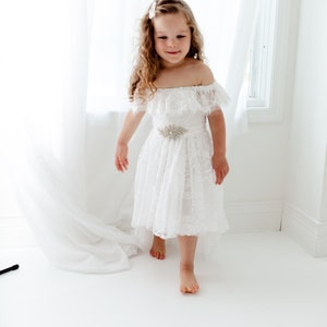 May include: A young girl wearing a white off-the-shoulder lace dress with a silver flower-shaped belt. She is standing on a white floor with a white curtain in the background.