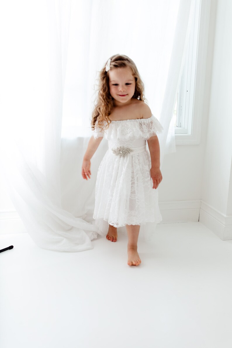 May include: A young girl wearing a white off-the-shoulder lace dress with a silver flower-shaped belt. She is standing on a white floor with a white curtain in the background.