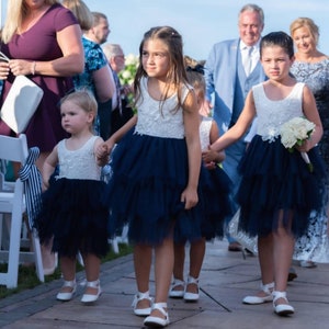 Puede incluir: Cuatro ni&ntilde;as peque&ntilde;as con vestidos blancos y azul marino caminan por el pasillo en una ceremonia de boda. Las ni&ntilde;as se toman de la mano y sonr&iacute;en. La ni&ntilde;a que va delante lleva un ramo de flores blancas.