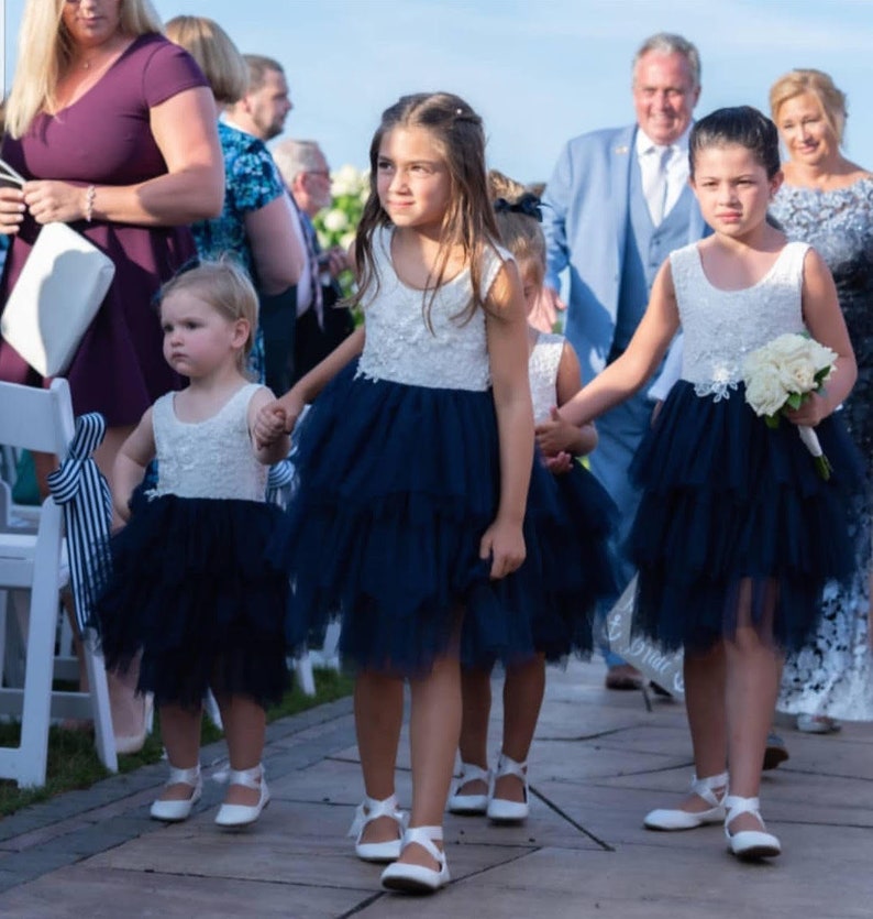 Puede incluir: Cuatro ni&ntilde;as peque&ntilde;as con vestidos blancos y azul marino caminan por el pasillo en una ceremonia de boda. Las ni&ntilde;as se toman de la mano y sonr&iacute;en. La ni&ntilde;a que va delante lleva un ramo de flores blancas.