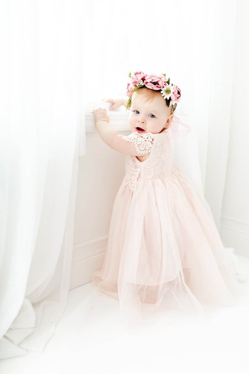 May include: A pale pink dress with a lace bodice and a layered tulle skirt, worn with a floral crown featuring pink and white flowers. The dress has short lace sleeves and a full, flowing skirt that reaches the floor, creating a soft appearance.
