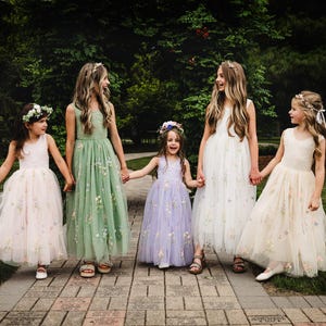 May include: Five children wearing floor-length tulle dresses in shades of pale pink, sage green, lavender, white, and cream. Each dress features delicate floral embroidery on the skirt and bodice, paired with matching floral headpieces in their hair.