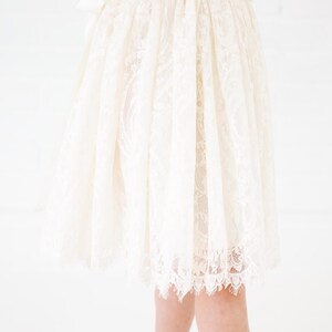 May include: A knee-length skirt crafted from cream-colored lace featuring an intricate floral pattern. The garment has a gathered waistline that creates a full, flowing silhouette, and the hem is finished with a delicate, scalloped lace edge.