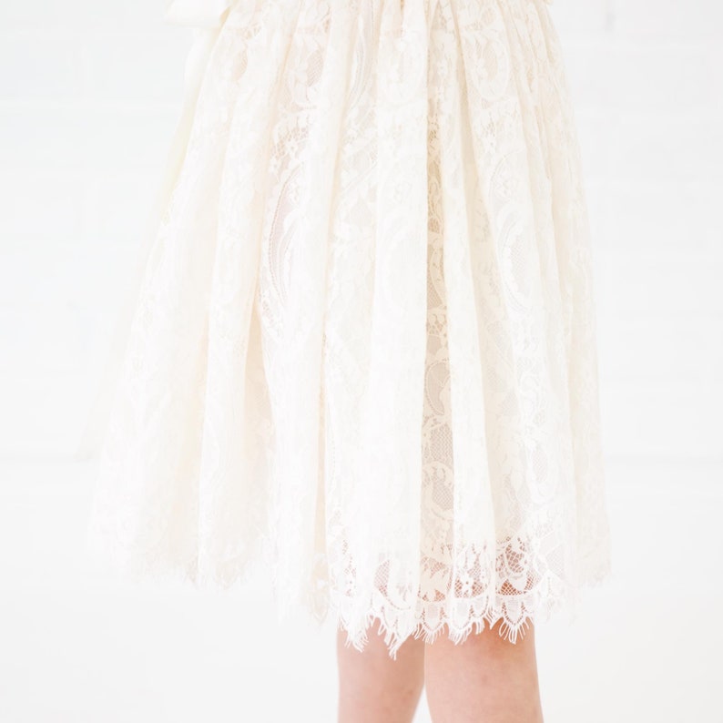 May include: A knee-length skirt crafted from cream-colored lace featuring an intricate floral pattern. The garment has a gathered waistline that creates a full, flowing silhouette, and the hem is finished with a delicate, scalloped lace edge.