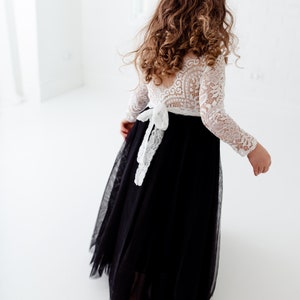 May include: A floor-length dress featuring a white long-sleeved lace bodice with an open back and a black tulle skirt. A white lace tie is fastened at the waist, creating a bow detail. The garment is shown from the back, highlighting the contrast.
