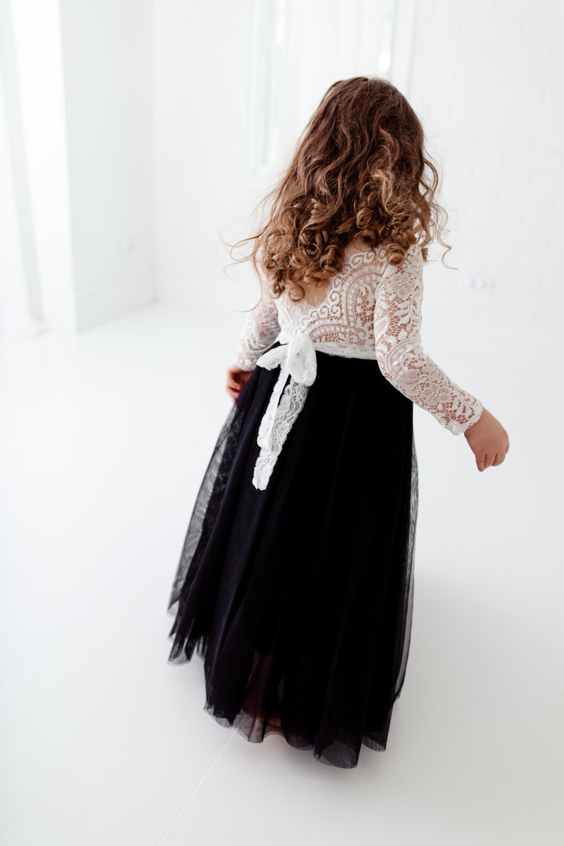May include: A floor-length dress featuring a white long-sleeved lace bodice with an open back and a black tulle skirt. A white lace tie is fastened at the waist, creating a bow detail. The garment is shown from the back, highlighting the contrast.