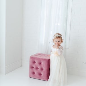 May include: A white dress with long lace sleeves, a scoop neckline, and a full tulle skirt featuring a large fabric flower at the waist. The dress is paired with a decorative headband with lace and bead details, standing beside a pink velvet ottoman.