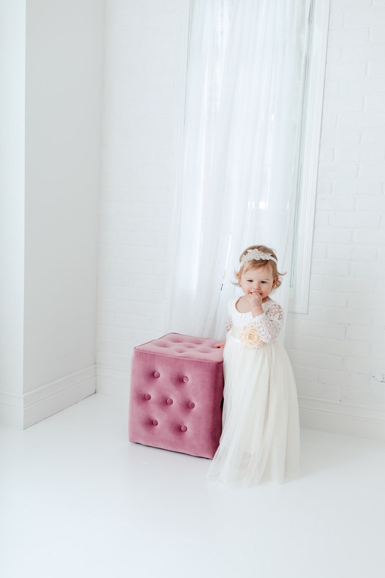 May include: A white dress with long lace sleeves, a scoop neckline, and a full tulle skirt featuring a large fabric flower at the waist. The dress is paired with a decorative headband with lace and bead details, standing beside a pink velvet ottoman.
