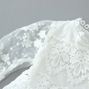 May include: White lace dress with long sleeves and a round neckline. The dress has a delicate floral pattern and is made of a soft, sheer fabric.