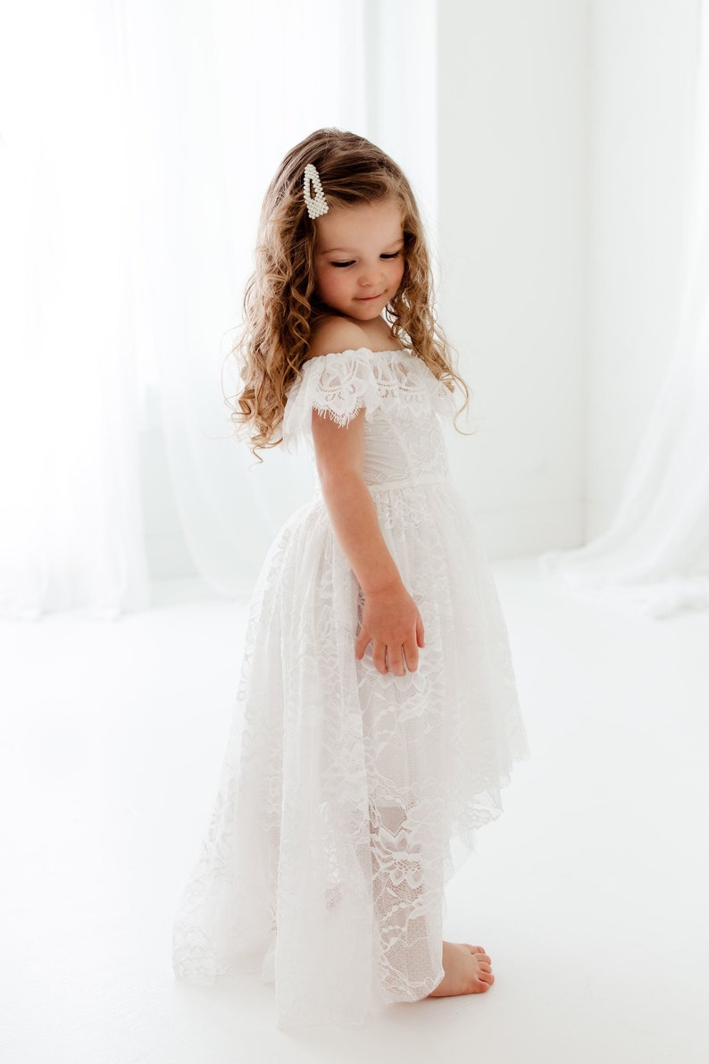 May include: A young girl wearing a white off-the-shoulder lace dress with a high-low hemline. She has curly brown hair and is wearing a pearl hair clip.