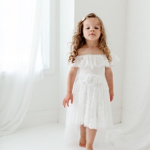May include: A young girl wearing a white off-the-shoulder lace dress with a flower accent on the waist. The dress has a high-low hemline and the girl is barefoot.
