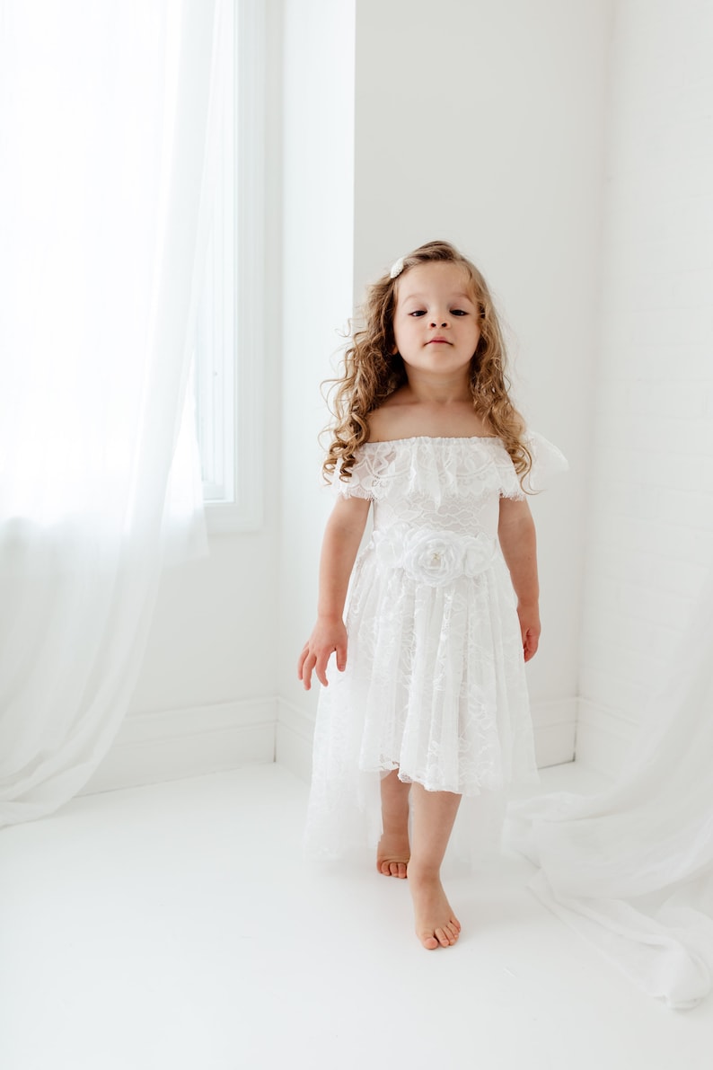 May include: A young girl wearing a white off-the-shoulder lace dress with a flower accent on the waist. The dress has a high-low hemline and the girl is barefoot.