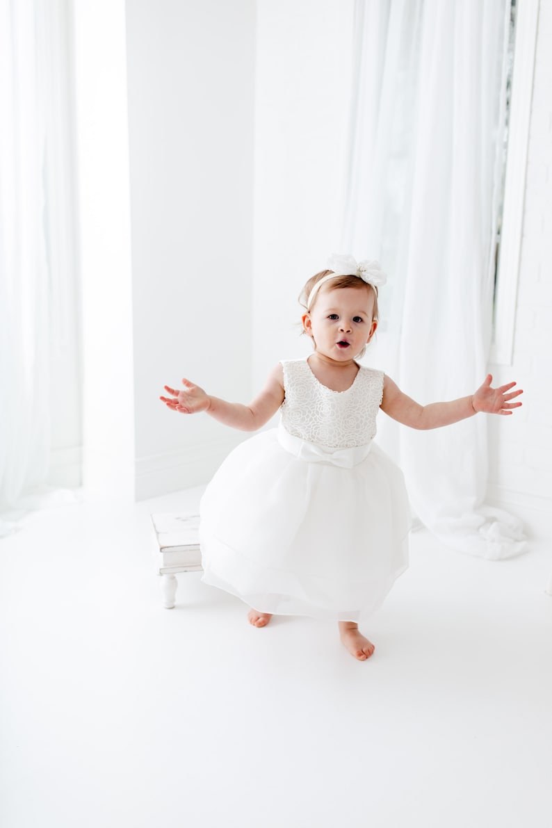 Puede incluir: Una ni&ntilde;a peque&ntilde;a con un vestido blanco de tul y una diadema de flores blancas. Est&aacute; de pie en una habitaci&oacute;n blanca con cortinas blancas.