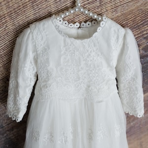 May include: White lace baby dress with long sleeves and a tulle skirt. The dress features a delicate floral pattern and a round neckline.