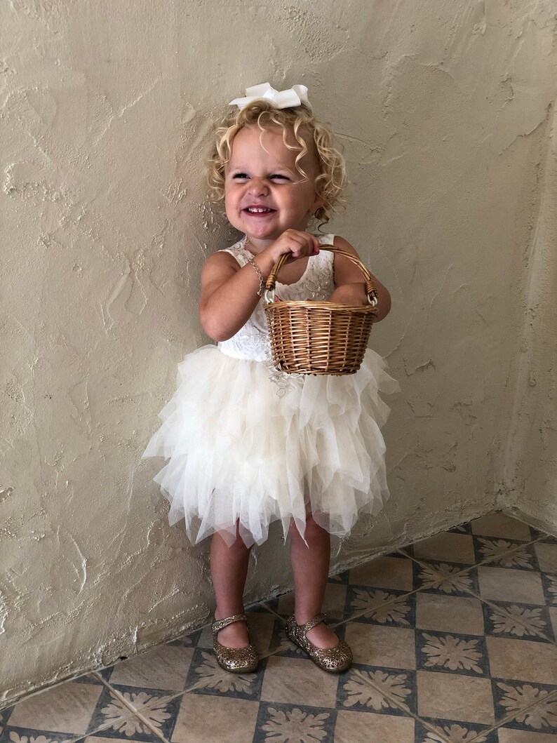 Op de afbeelding: Een jong meisje in een witte tule jurk en een witte strik in haar haar houdt een rieten mandje vast. Ze glimlacht en kijkt naar de zijkant. Het meisje staat op een betegelde vloer.