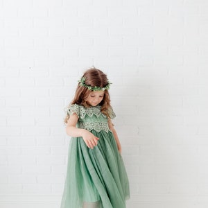 May include: A young girl wearing a sage green tulle dress with a lace bodice and a leafy green crown. The dress has a flowing skirt and the girl is standing on a white background.